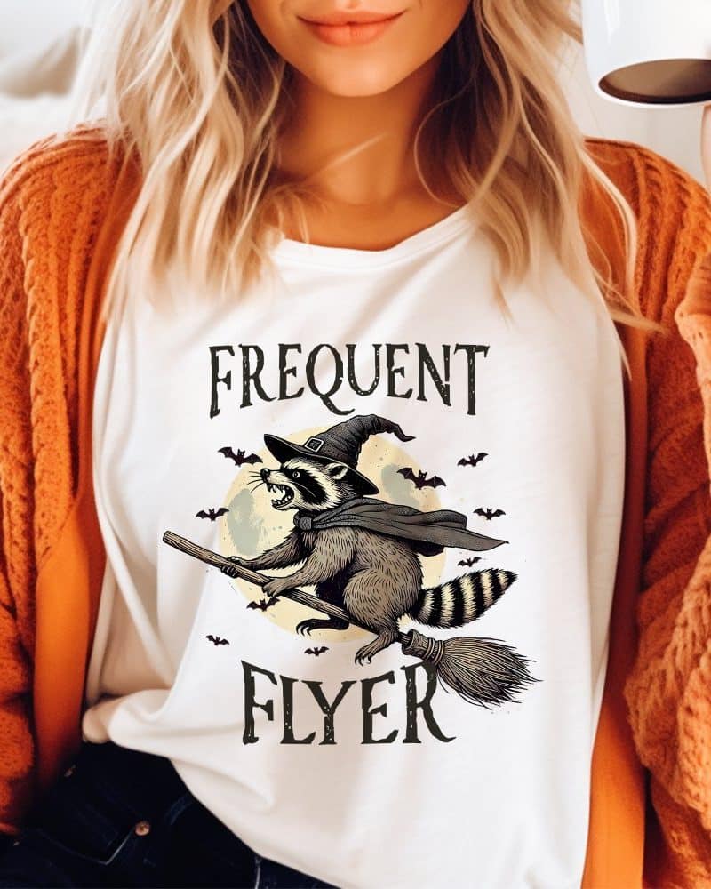 White shirt with a graphic of a raccoon wearing a witch's hat and flying on a broomstick in front of a full moon. The text "Frequent Flyer" is above and below the image.