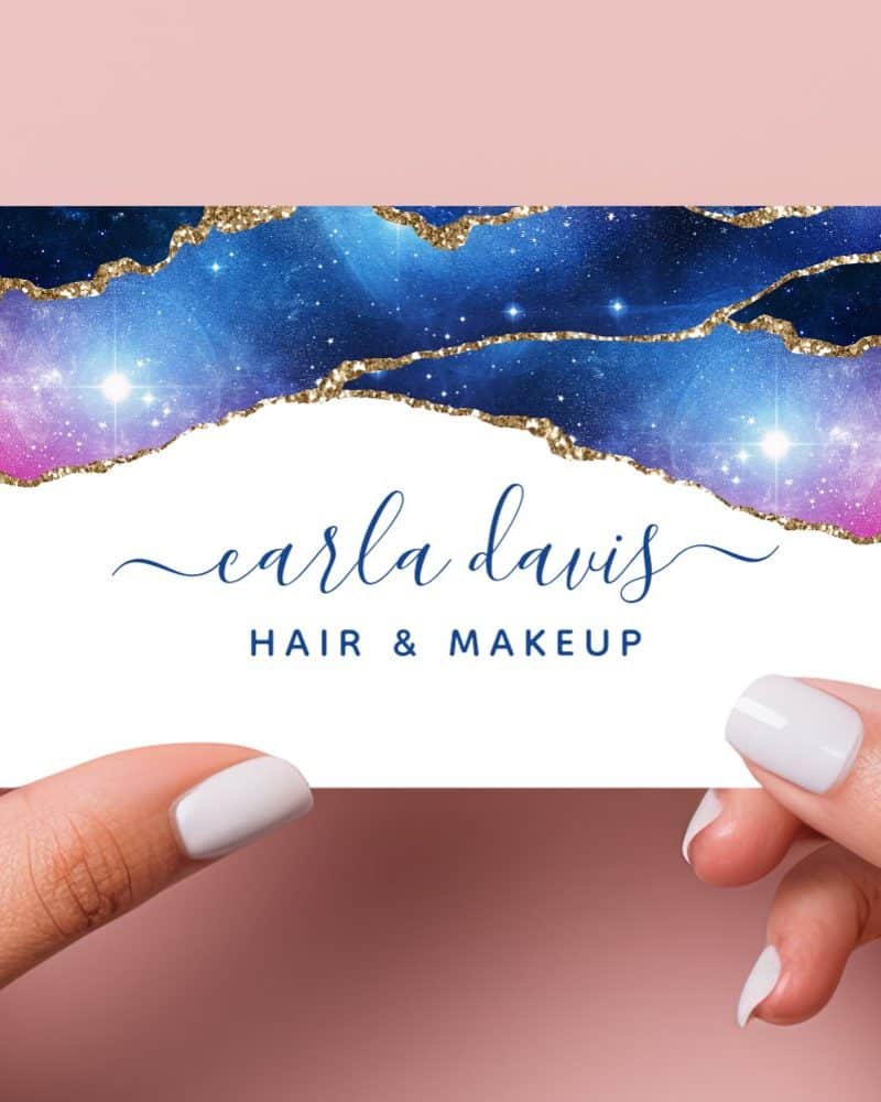 Two hands hold up a business card featuring a galaxy-themed design with gold glitter accents. The card displays Carla Davis Hair & Makeup.