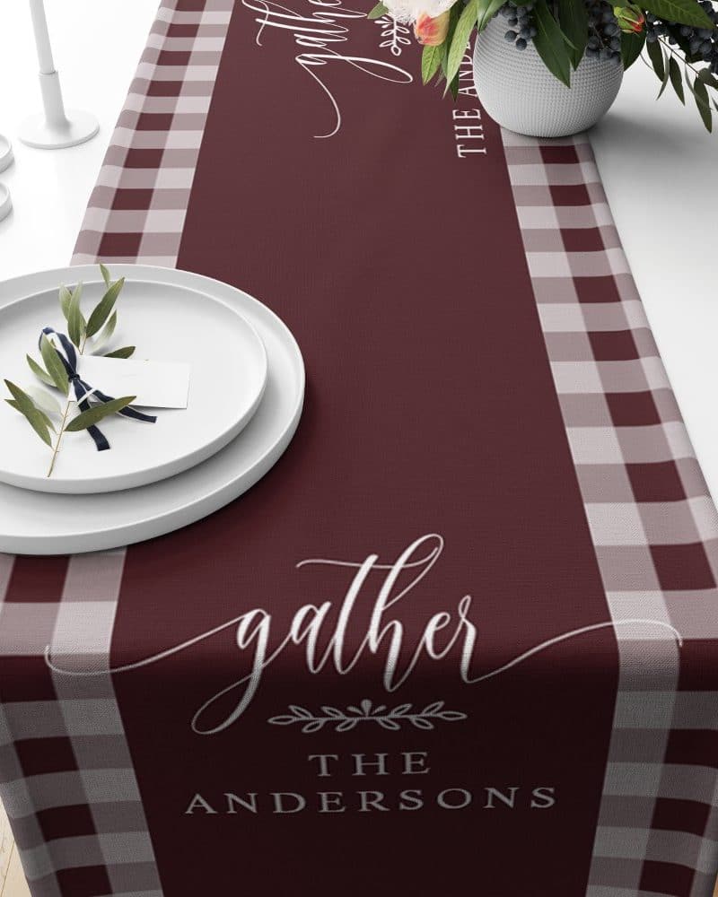 Burgundy table runner with gray and white gingham borders, personalized with the word gather and the family name Andersons.