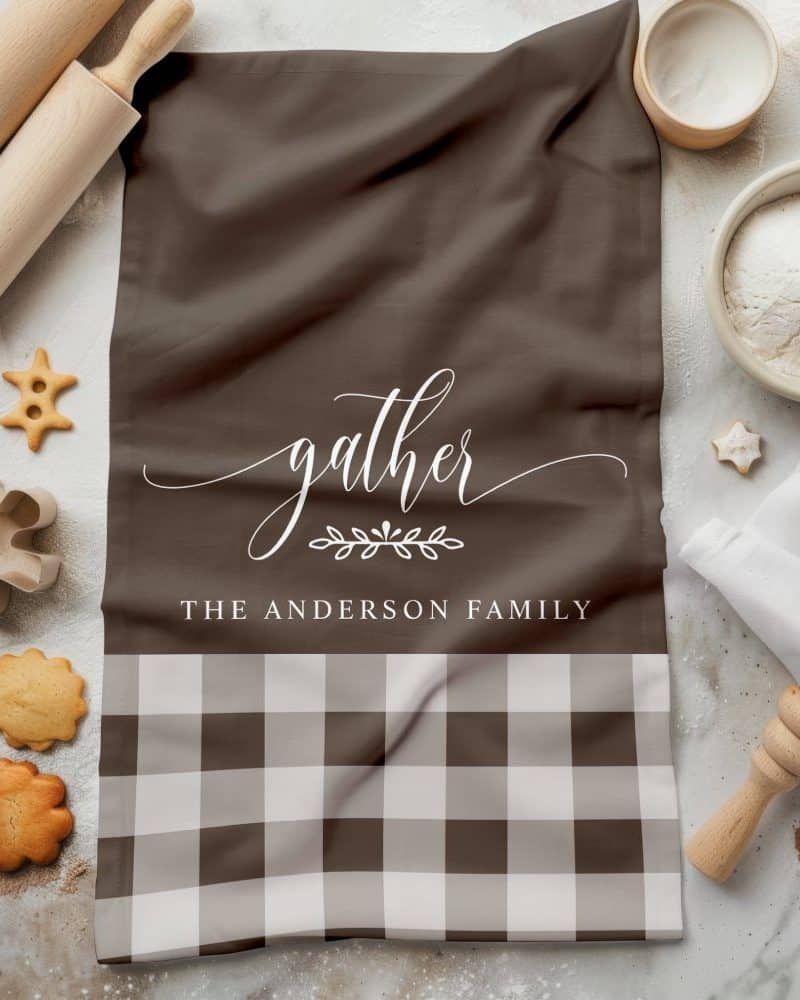 Brown kitchen towel with white script reading Gather, The Anderson Family, and a brown and white gingham bottom border.