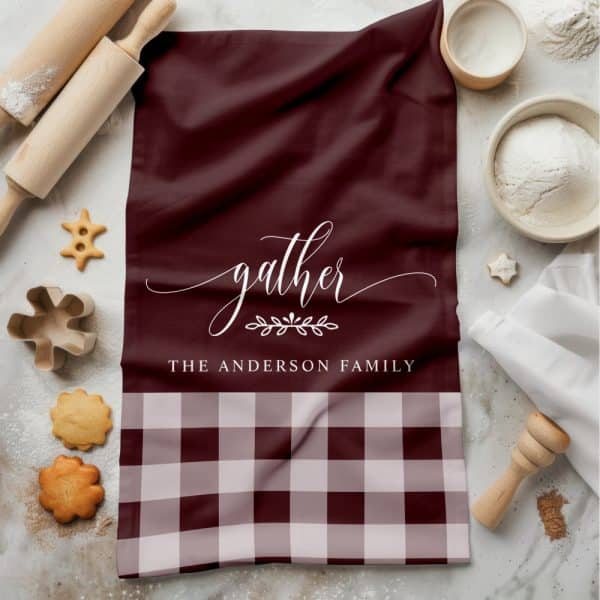 A Maroon Kitchen Towel With The Word Gather In A Script Font, A Small Leaf Design, And The Anderson Family Printed Below. The Bottom Of The Towel Is A Maroon And Cream Gingham Pattern.