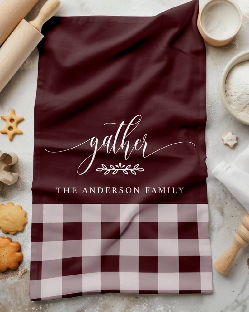 A maroon kitchen towel with the word gather in a script font, a small leaf design, and The Anderson Family printed below. The bottom of the towel is a maroon and cream gingham pattern.