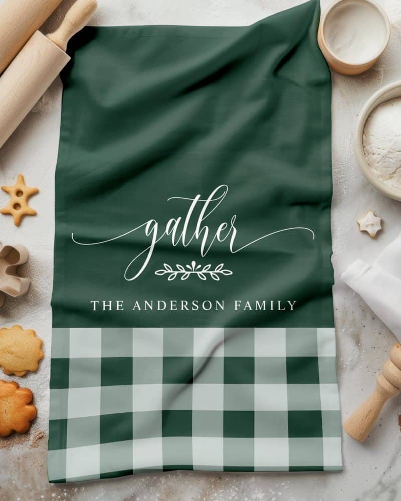Green tea towel with the word gather and The Anderson Family in white lettering. The bottom features a green and white gingham pattern.