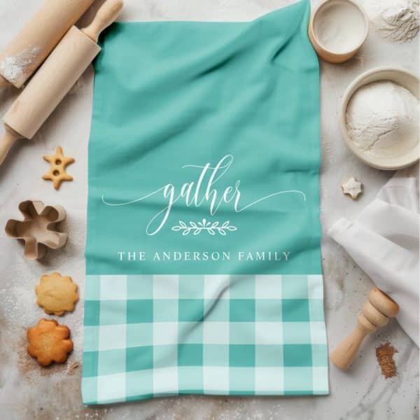 Teal Kitchen Towel With The Word Gather In A Script Font, A Small Floral Design, And The Anderson Family Printed Below. The Bottom Half Of The Towel Is Teal And White Gingham.