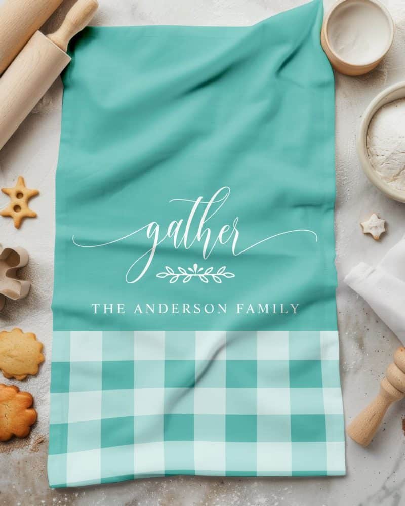 Teal kitchen towel with the word gather in a script font, a small floral design, and The Anderson Family printed below. The bottom half of the towel is teal and white gingham.