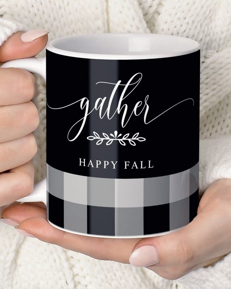 A person's hands holding a black coffee mug. The mug features the word gather in elegant script, a small leaf design, and Happy Fall below, all in white against a black and gray buffalo plaid background.