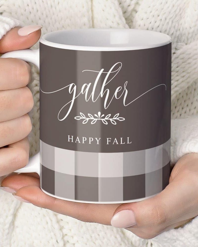Two hands hold a coffee mug. The mug is dark brown with a light gray and dark brown gingham pattern at the bottom and the words Gather and Happy Fall in a script font.
