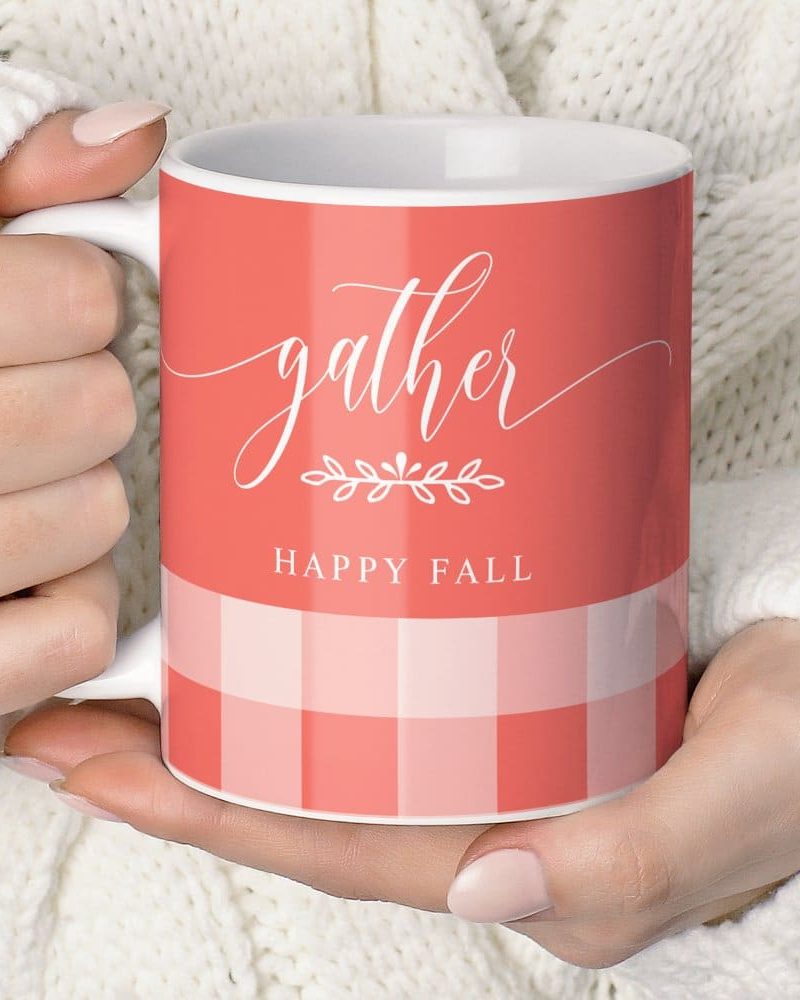 A hand holds a coral-colored mug with the words Gather and Happy Fall. The mug has a gingham pattern at the bottom.