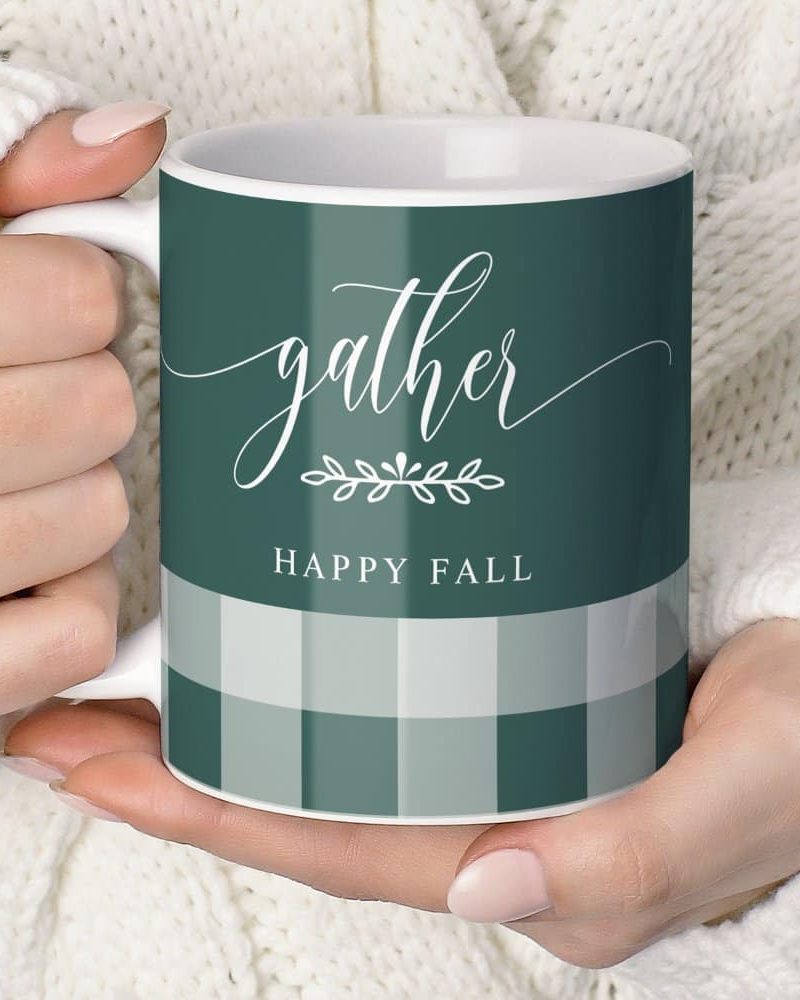 A person's hands holding a teal mug with the words Gather and Happy Fall. A simple leaf design is between the words. The mug also features a subtle green and white gingham pattern.