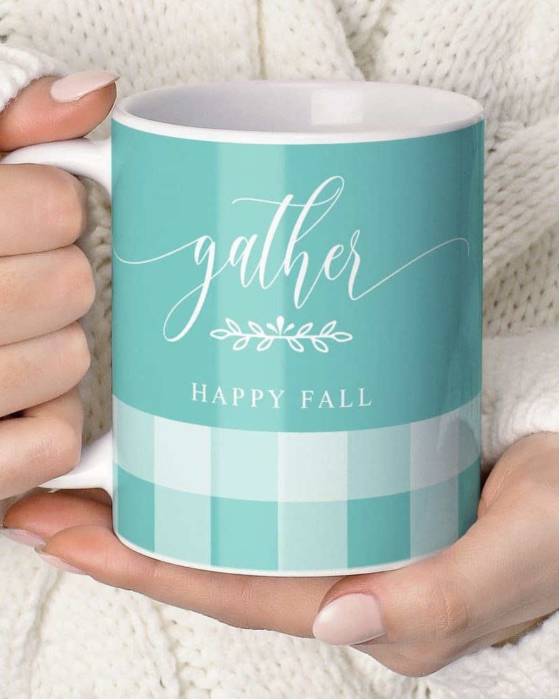 A teal mug with the words Gather and Happy Fall, decorated with a small leaf design and a gingham pattern, held in a person's hands.