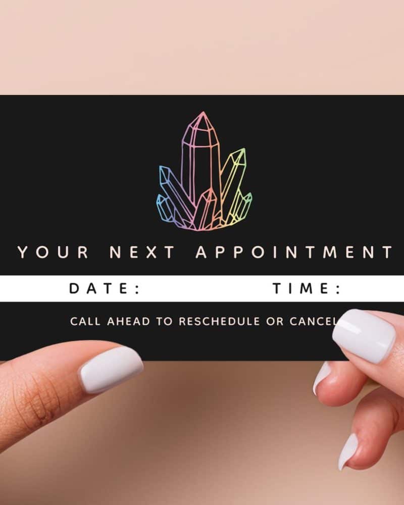 Two hands hold a black appointment card featuring a pastel crystal illustration. The card displays the text "Your Next Appointment," with lines for date and time. A note indicates to call ahead to reschedule or cancel.