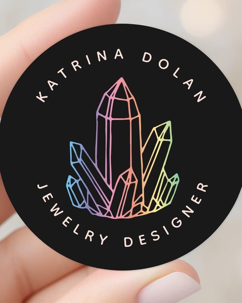 Black circle sticker with a rainbow crystal cluster graphic and the text Katrina Dolan Jewelry Designer.