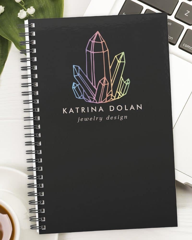 Black spiral notebook with a rainbow crystal logo and Katrina Dolan jewelry design text.