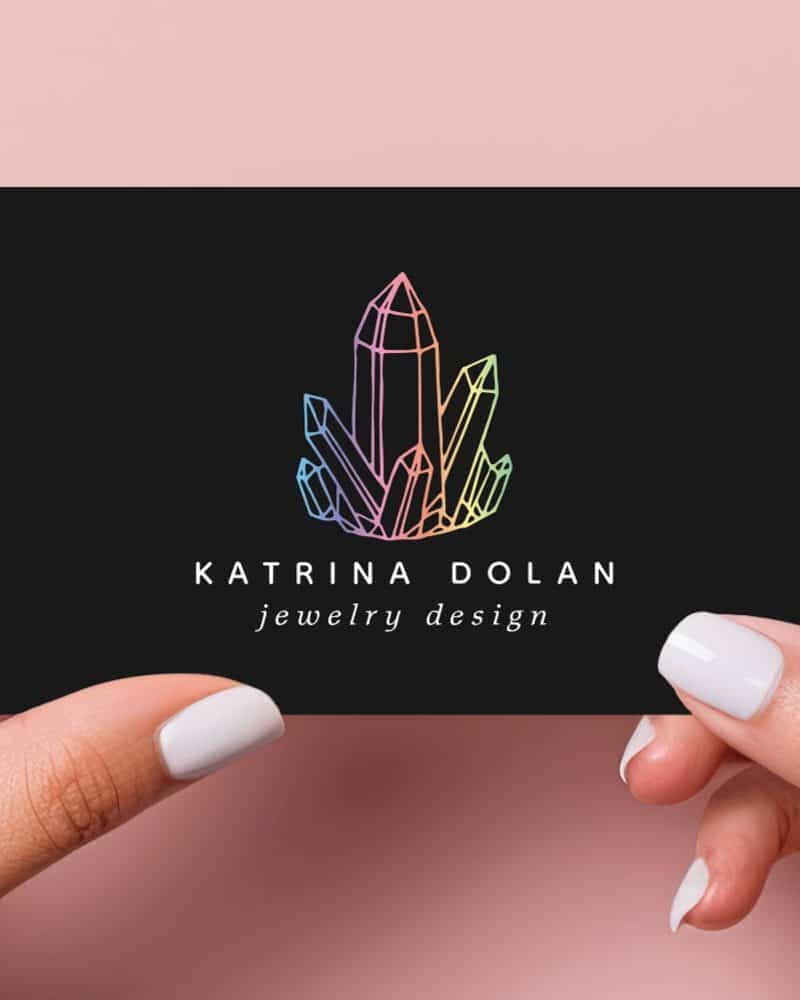 Two hands hold a black business card featuring a pastel rainbow crystal cluster logo and the text Katrina Dolan jewelry design.