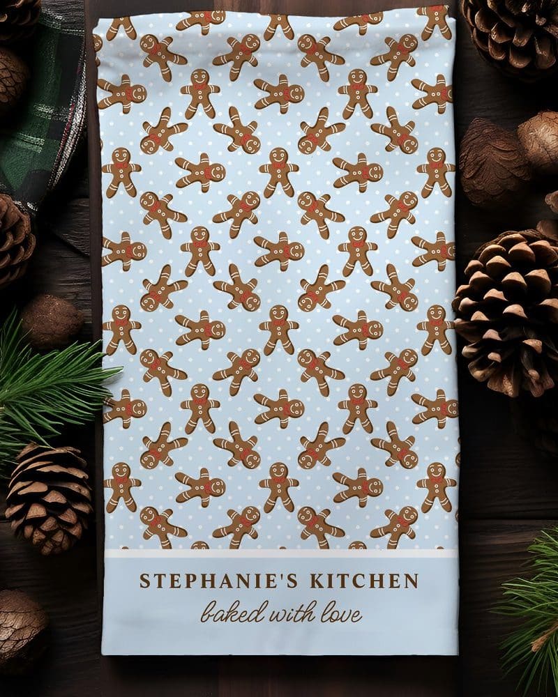 A light blue kitchen towel with a repeating pattern of gingerbread men, personalized with the text Stephanie's Kitchen baked with love.