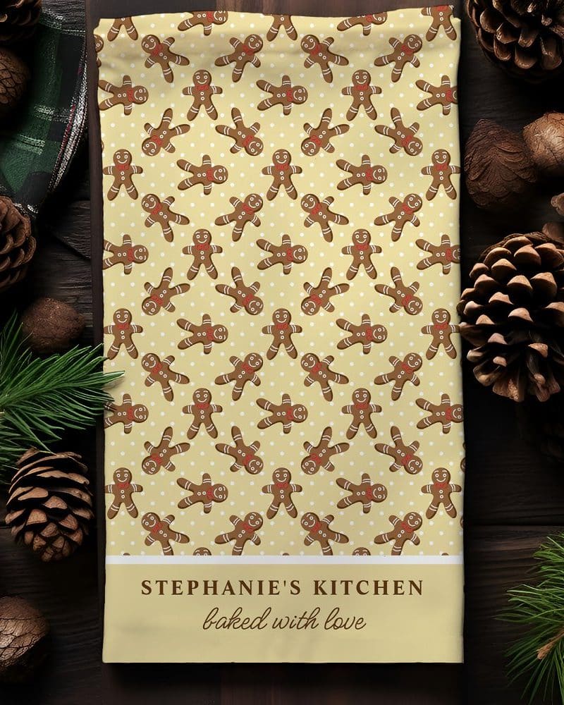 A kitchen tea towel featuring a repeating pattern of gingerbread men on a pale yellow background. The bottom of the towel is personalized with Stephanie's Kitchen baked with love.