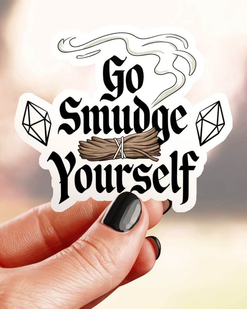 Hand holding a sticker that says Go Smudge Yourself in stylized text, with an illustration of smudge sticks and crystals.
