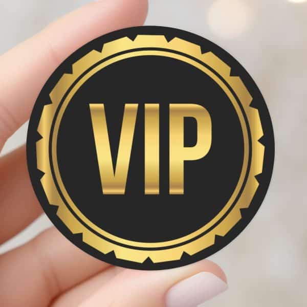 A Hand Holding A Round Vip Sticker With Gold Lettering On A Black Background.