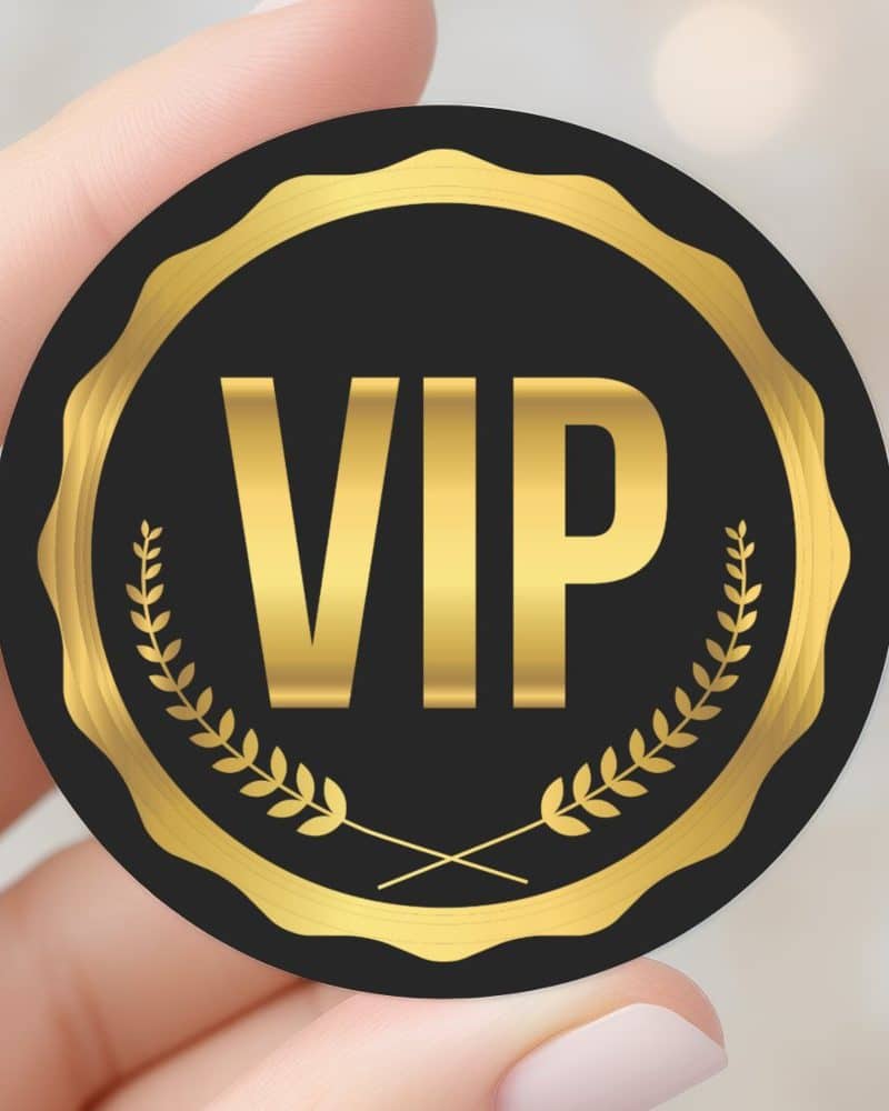 A hand holding a black VIP sticker with gold lettering and laurel wreaths.