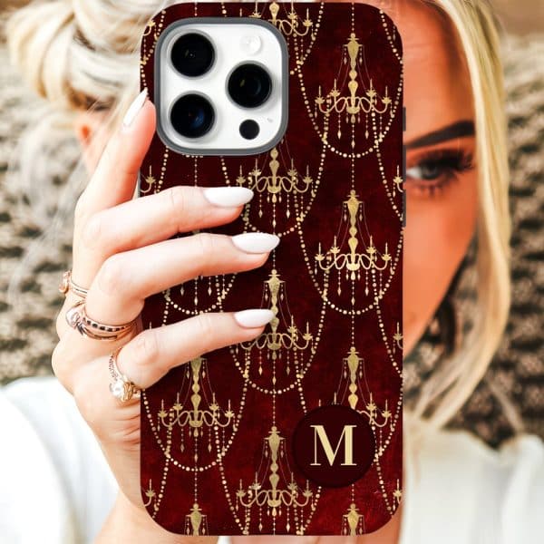 A Woman'S Hand Holds A Phone Case With A Burgundy Background And A Repeating Pattern Of Gold Chandeliers. A Gold Initial M Is Centered On The Case.