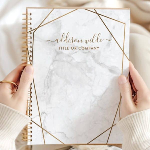 Two Hands Hold A Spiral-Bound Notebook With A Marble Cover And Gold Geometric Design. The Name Addison Wilde Is Printed On The Cover.