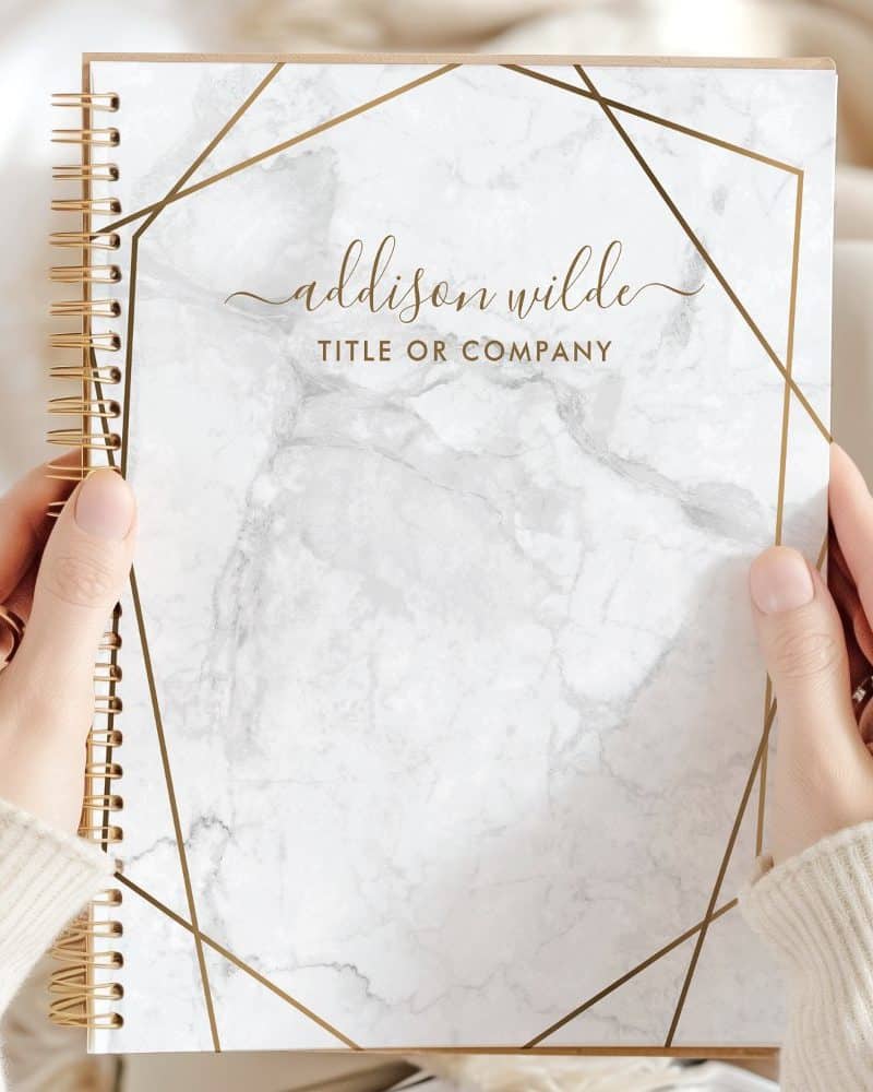 Two hands hold a spiral-bound notebook with a marble cover and gold geometric design. The name Addison Wilde is printed on the cover.