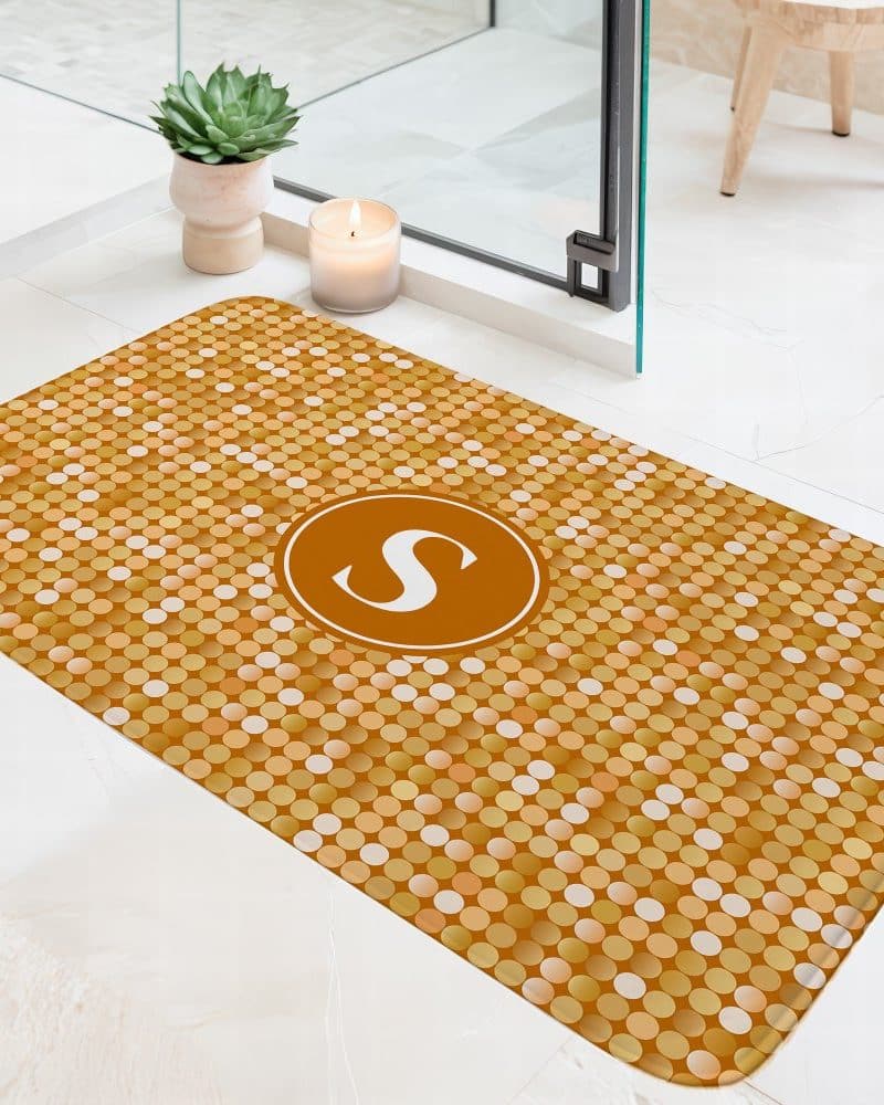 Gold bath mat with a pattern of small circles and the letter S in a circular emblem in the center.