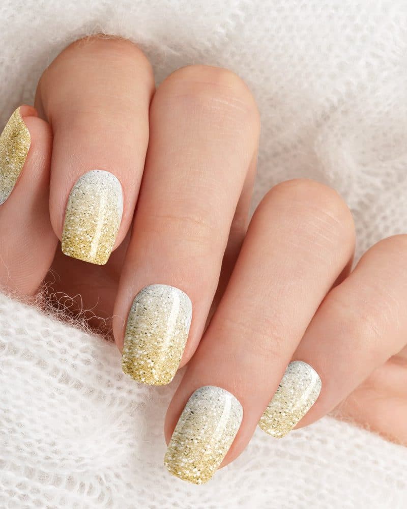 Close-up of a hand with fingernails painted in a gold and white glitter ombre design.