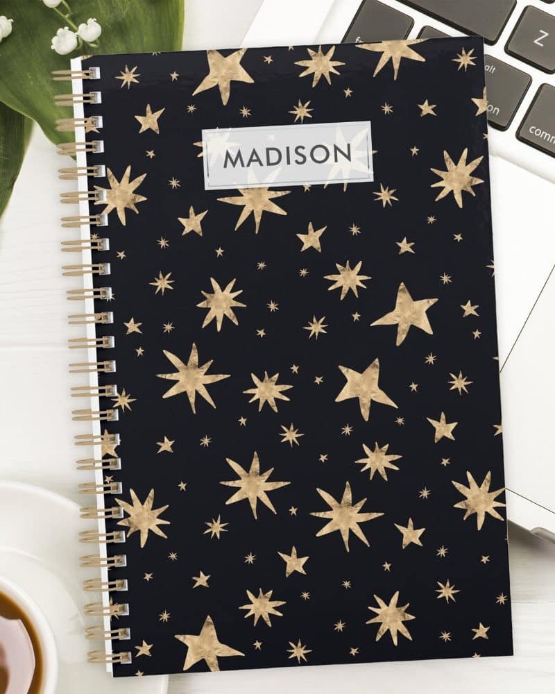 Spiral-bound notebook with a dark background and gold star pattern; the name Madison is printed on the cover.