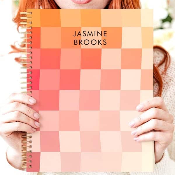 Person Holding A Spiral-Bound Notebook With A Peach And Coral Pixelated Cover. The Name Jasmine Brooks Is Printed On The Cover.