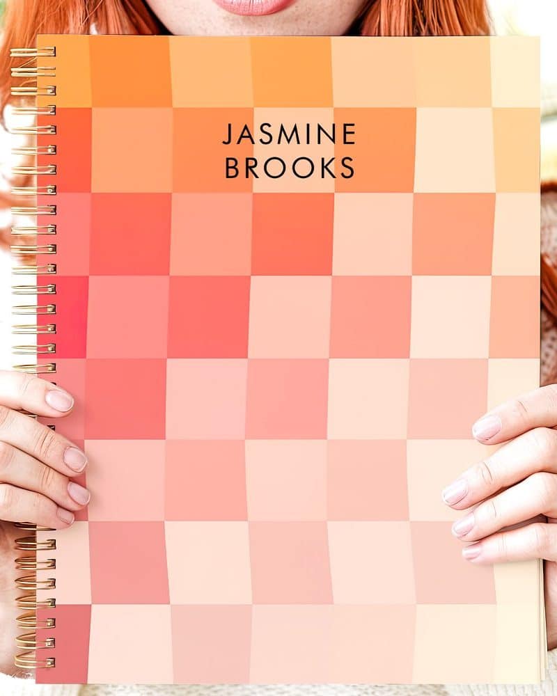 Person holding a spiral-bound notebook with a peach and coral pixelated cover. The name Jasmine Brooks is printed on the cover.