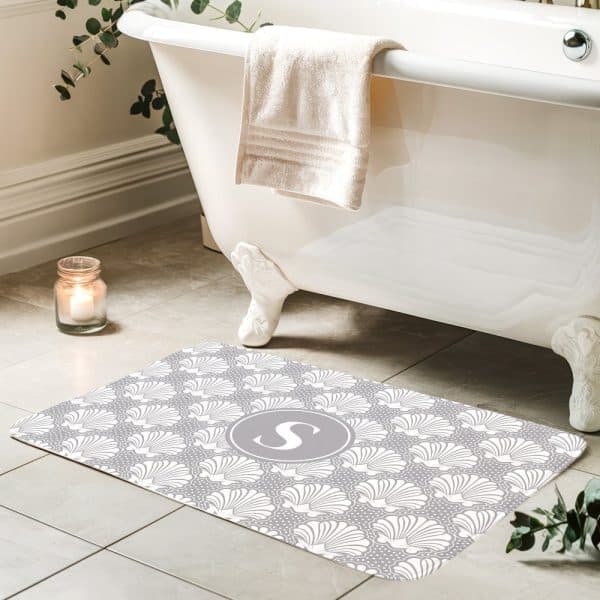 Gray Bath Mat With A White Seashell Pattern And A Gray Circle Monogram In The Center.