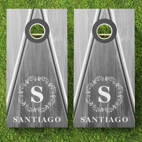 Pair Of Gray Personalized Cornhole Boards. Each Board Features A Gray Wood Grain Design, A Monogrammed Wreath, And The Name Santiago.