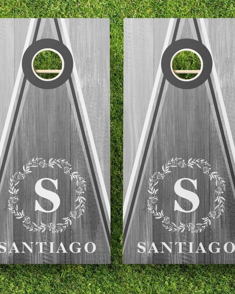 Pair of gray personalized cornhole boards. Each board features a gray wood grain design, a monogrammed wreath, and the name Santiago.