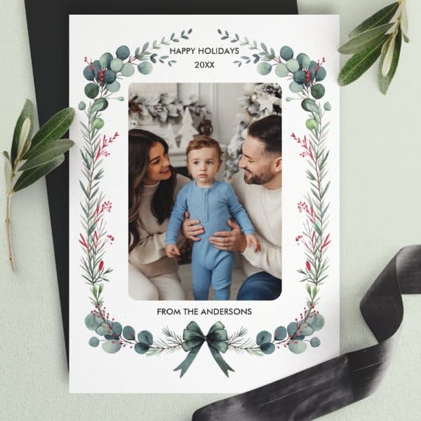 Holiday Photo Card Featuring A Family Of Three, Parents And A Toddler, Within A Greenery Frame.