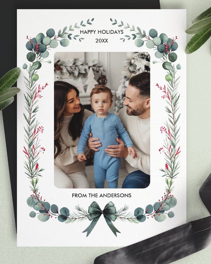 Holiday photo card featuring a family of three, parents and a toddler, within a greenery frame.