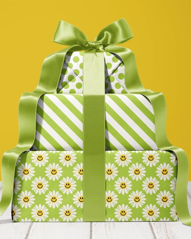 A stack of three gifts wrapped in green and white paper, tied with a large green ribbon. The bottom gift features a daisy and smiley face pattern.