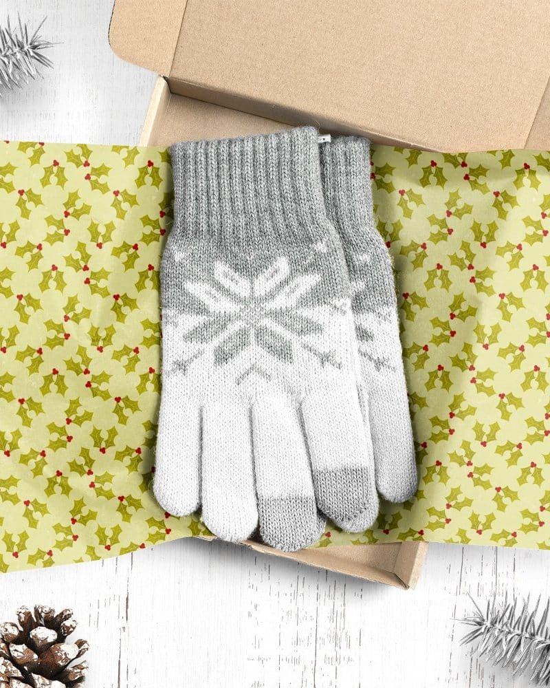 Pair of gray and white knit gloves with snowflake pattern, presented in a gift box lined with holly-patterned tissue paper.