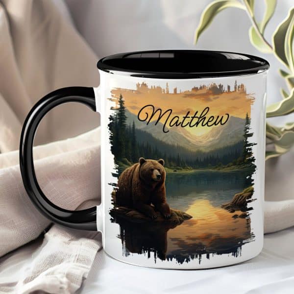 A Mug With A Black Handle And Interior, Featuring A Grizzly Bear Sitting By A Lake At Sunset In A Mountain Landscape. The Name Matthew Is Printed Above The Scene.