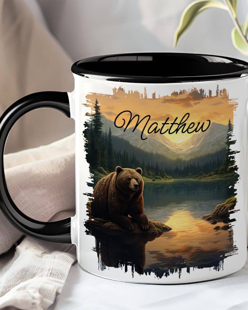 A mug with a black handle and interior, featuring a grizzly bear sitting by a lake at sunset in a mountain landscape. The name Matthew is printed above the scene.
