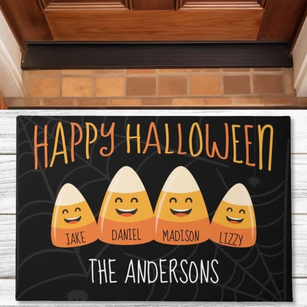 A Black Halloween Doormat With The Text Happy Halloween, Three Smiling Candy Corn Characters With The Names Jake, Daniel, And Lizzy, And The Family Name The Andersons.