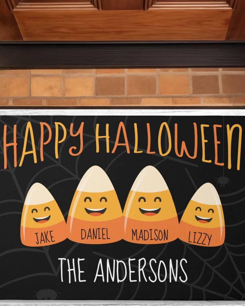 A black Halloween doormat with the text Happy Halloween, three smiling candy corn characters with the names Jake, Daniel, and Lizzy, and the family name The Andersons.