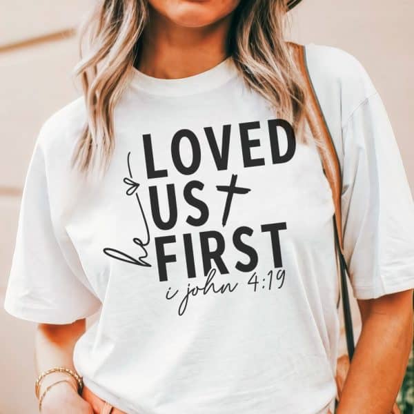 White T-Shirt With Black Lettering That Says He Loved Us First, And 1 John 4:19 Below.