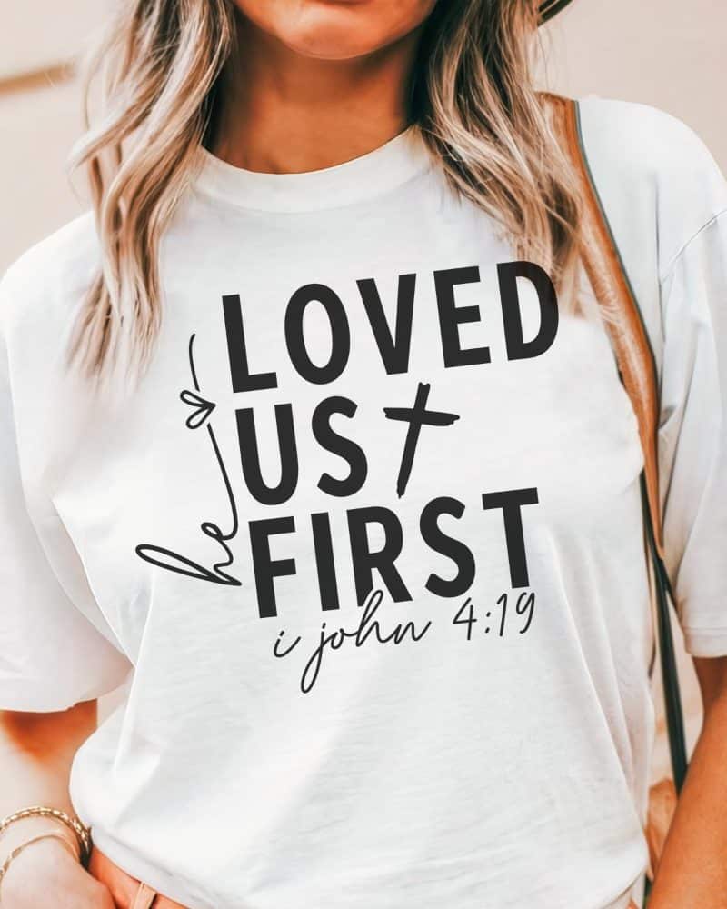 White t-shirt with black lettering that says He Loved Us First, and 1 John 4:19 below.