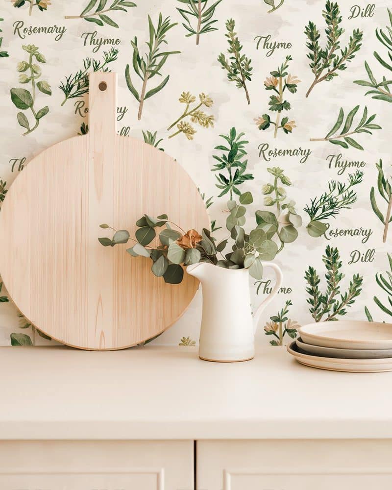 A round wooden cutting board and a small white creamer sit on a light beige countertop in front of a wall featuring herb-themed wallpaper.