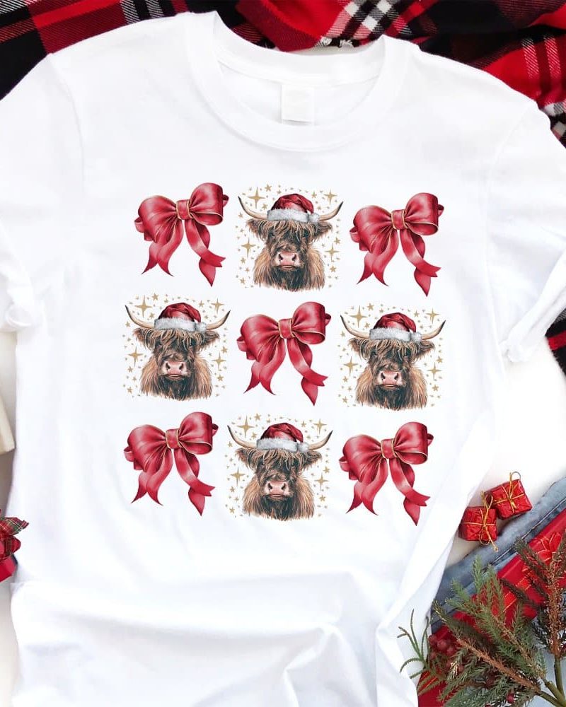 White t-shirt with a repeating pattern of Highland cows wearing Santa hats and red bows.