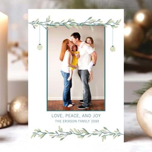 Christmas Card Featuring A Family Of Four; Parents Kissing With Their Two Young Children. A Simple Leafy Border And Ornaments Frame The Photo.