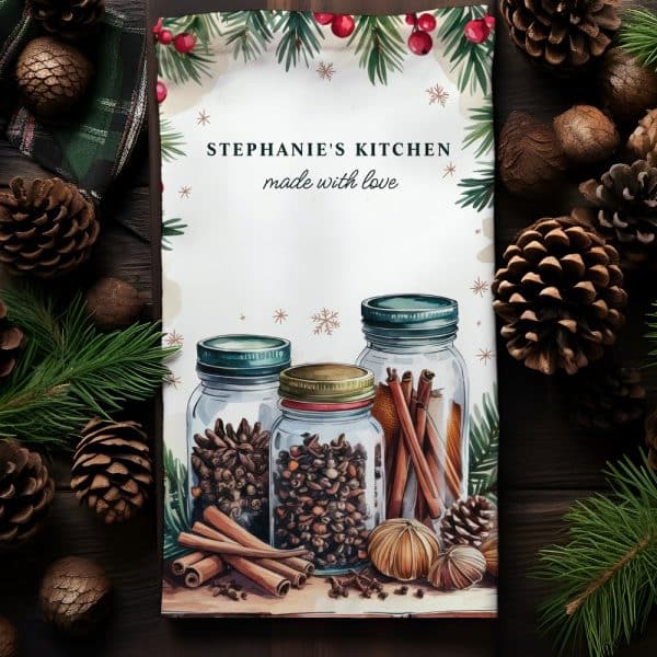 A Kitchen Towel Featuring Three Glass Jars Filled With Spices And Cinnamon Sticks, Surrounded By Pine Branches And Pine Cones. The Towel Is Personalized With The Text Stephanie'S Kitchen, Made With Love.
