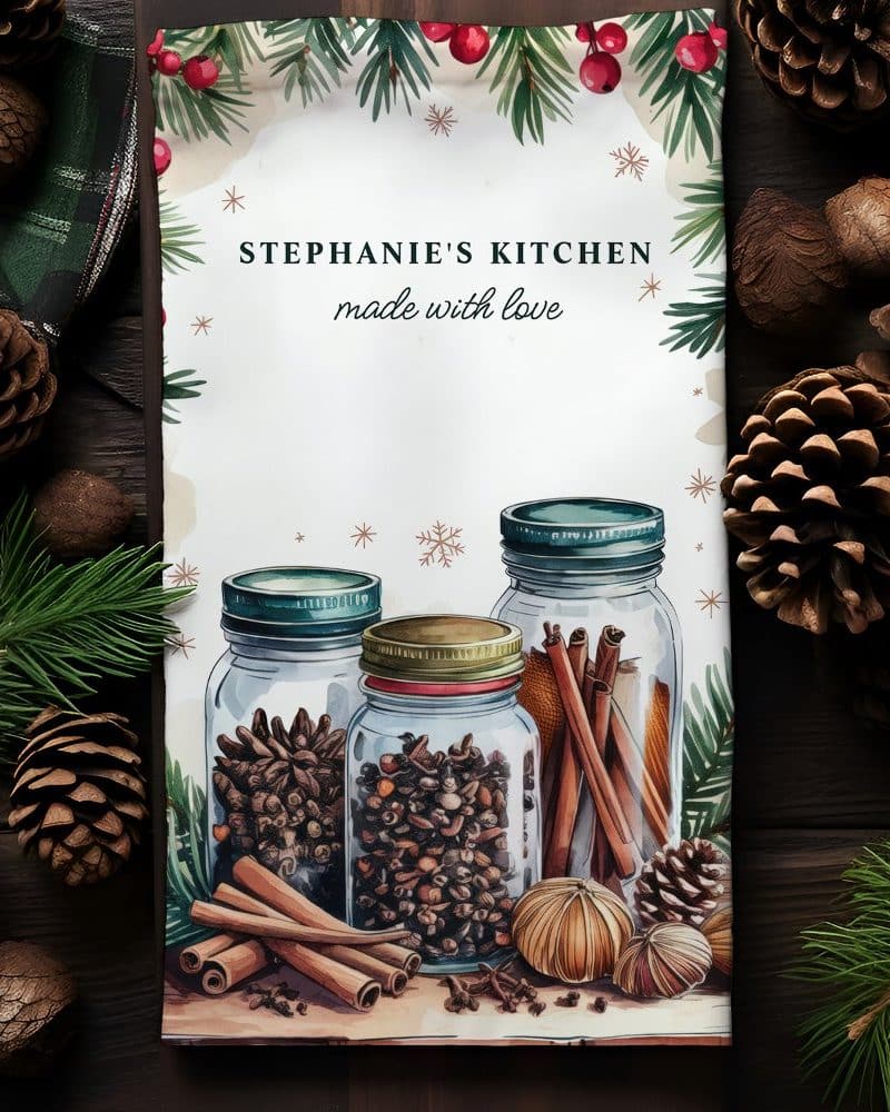 A kitchen towel featuring three glass jars filled with spices and cinnamon sticks, surrounded by pine branches and pine cones. The towel is personalized with the text Stephanie's Kitchen, made with love.