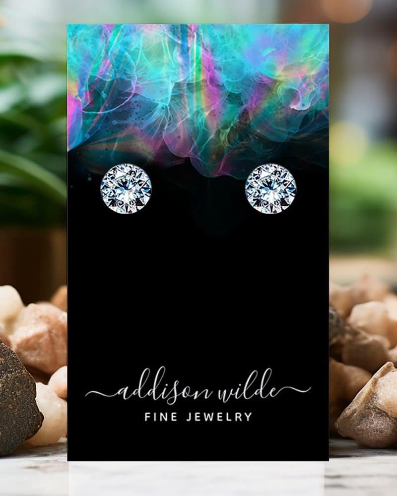 Pair of round diamond stud earrings displayed on a black card with a colorful background. The card includes the text Addison Wilde Fine Jewelry.